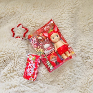 May include: A clear plastic package containing a small doll wearing a strawberry-themed outfit, a Hello Kitty figurine, and a box of Pocky. A red strawberry-shaped keychain and a small red and white package with Japanese text are also visible.