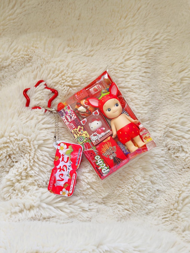 May include: A clear plastic package containing a small doll wearing a strawberry-themed outfit, a Hello Kitty figurine, and a box of Pocky. A red strawberry-shaped keychain and a small red and white package with Japanese text are also visible.