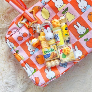 Peut inclure: Un sac fourre-tout &agrave; carreaux roses et orange avec des illustrations de Miffy et de fruits. Une pochette en plastique transparent est fix&eacute;e &agrave; la bandouli&egrave;re, contenant des jouets miniatures, dont des paquets BelVita, Jell-O et Wet Ones.