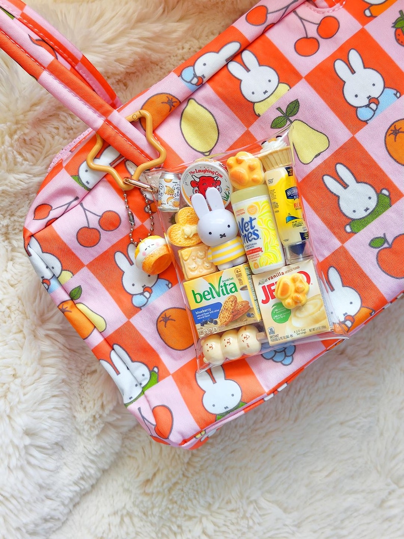 Peut inclure: Un sac fourre-tout &agrave; carreaux roses et orange avec des illustrations de Miffy et de fruits. Une pochette en plastique transparent est fix&eacute;e &agrave; la bandouli&egrave;re, contenant des jouets miniatures, dont des paquets BelVita, Jell-O et Wet Ones.