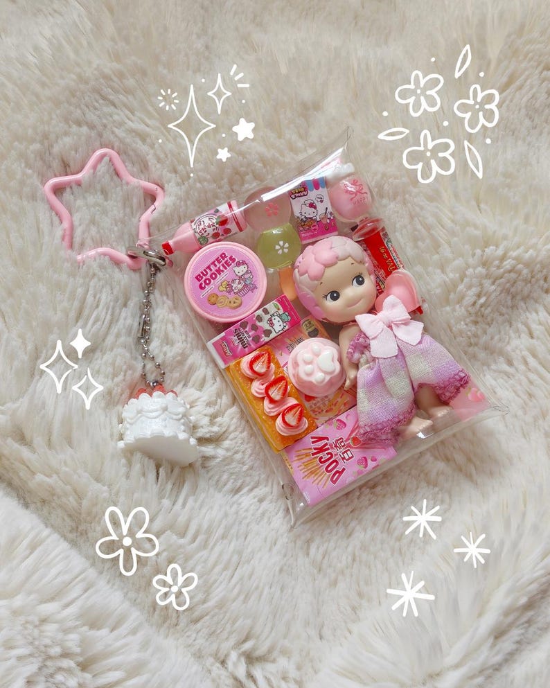 K&ouml;nnte beinhalten: Eine transparente Plastikt&uuml;te gef&uuml;llt mit Miniatur-S&uuml;&szlig;igkeiten in Rosa und Wei&szlig;, einer Puppe und Lipgloss. Die Tasche ist an einem rosa sternf&ouml;rmigen Schl&uuml;sselanh&auml;nger und einem kleinen wei&szlig;en Kuchenanh&auml;nger befestigt. Die Artikel sind auf einer flauschigen wei&szlig;en Oberfl&auml;che angeordnet.
