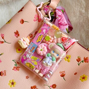 May include: A pink leather handbag with a floral pattern, holding a clear pouch filled with candies and a small doll. The pouch contains a variety of colorful sweets and a Hello Kitty lip gloss. A pink star-shaped keychain and a floral scarf are attached.