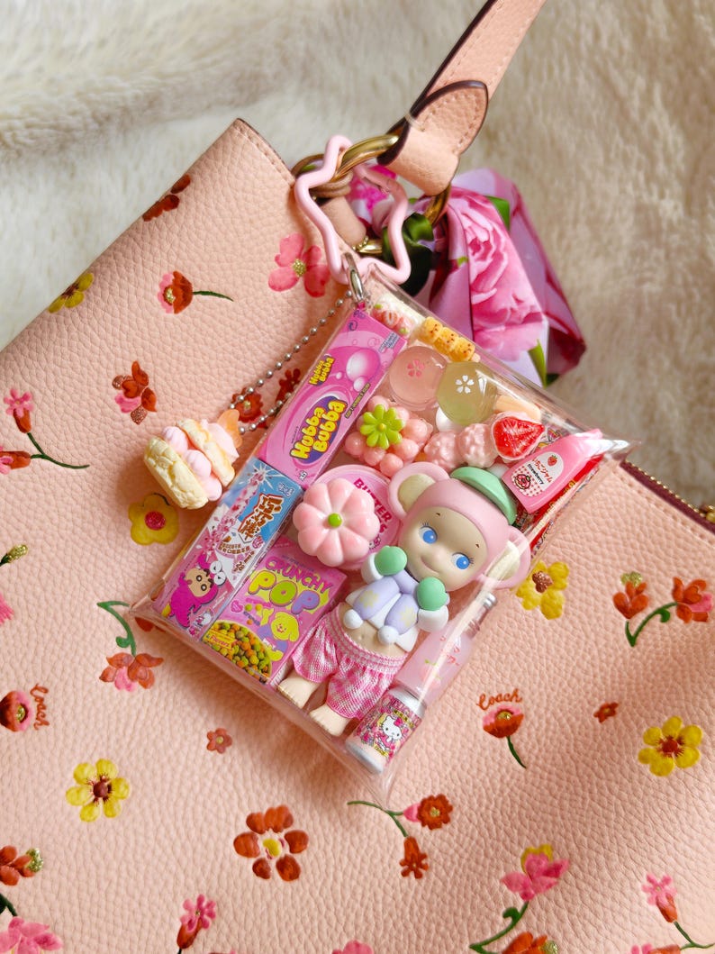 May include: A pink leather handbag with a floral pattern, holding a clear pouch filled with candies and a small doll. The pouch contains a variety of colorful sweets and a Hello Kitty lip gloss. A pink star-shaped keychain and a floral scarf are attached.