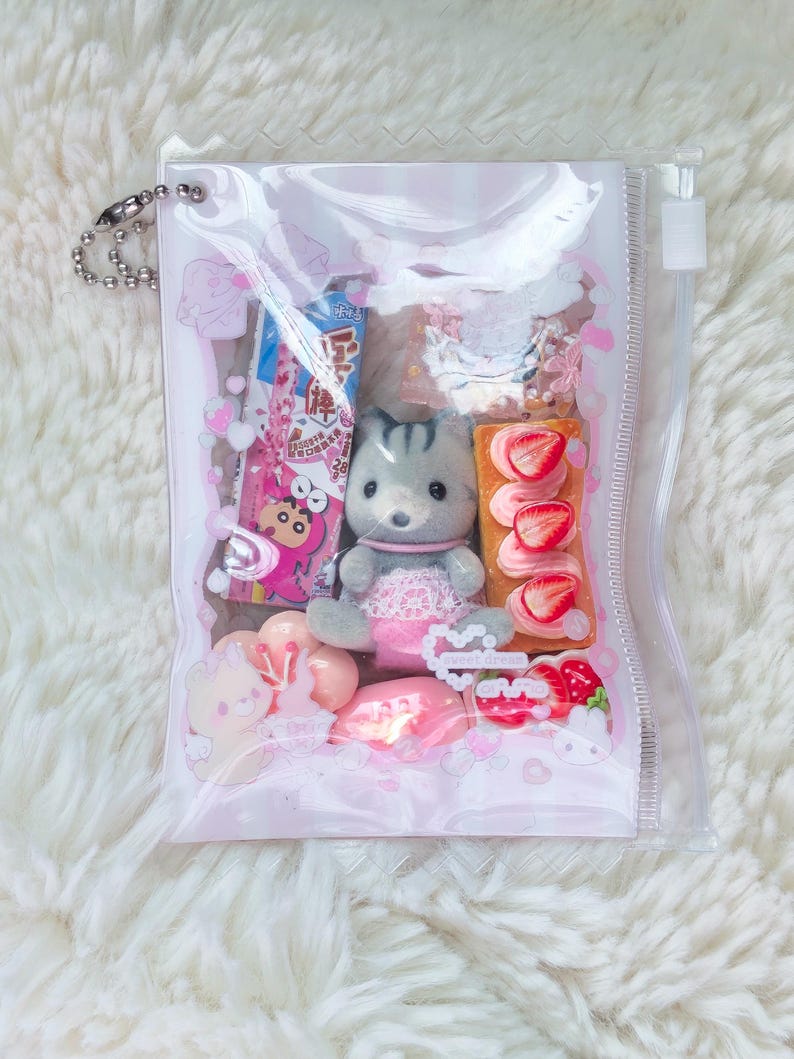 K&ouml;nnte beinhalten: Eine transparente Plastikt&uuml;te gef&uuml;llt mit Miniaturspielzeug und Snacks. Die T&uuml;te enth&auml;lt eine graue Katzenfigur, einen kleinen Kuchen mit Erdbeeren und verschiedene rosa und wei&szlig;e S&uuml;&szlig;igkeiten. Oben ist eine silberne Kette befestigt.