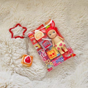 May include: A clear plastic bag containing a small doll wearing a strawberry hat and shorts with yellow hearts. The bag also contains miniature food items and is attached to a red star-shaped keychain and a charm shaped like a piece of toast with a strawberry on top.