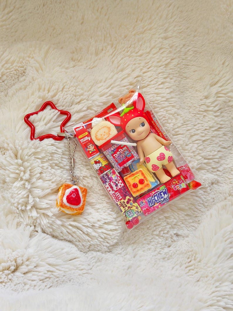 May include: A clear plastic bag containing a small doll wearing a strawberry hat and shorts with yellow hearts. The bag also contains miniature food items and is attached to a red star-shaped keychain and a charm shaped like a piece of toast with a strawberry on top.