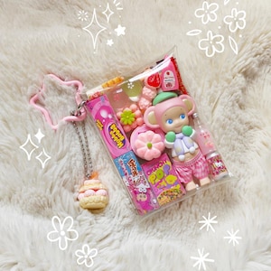 May include: A clear plastic pouch filled with colorful candies, a small doll, and a miniature bottle. The pouch is attached to a pink star-shaped keychain and a cupcake charm. The items are arranged on a fluffy white surface.