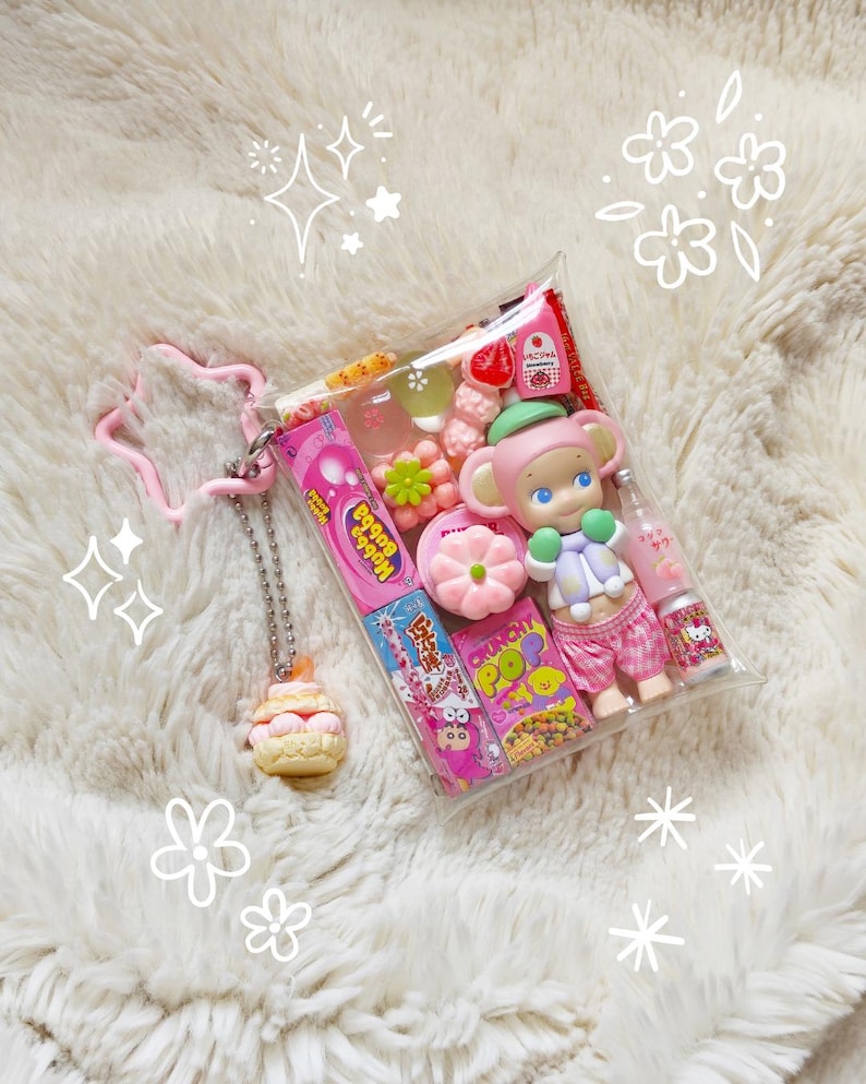 May include: A clear plastic pouch filled with colorful candies, a small doll, and a miniature bottle. The pouch is attached to a pink star-shaped keychain and a cupcake charm. The items are arranged on a fluffy white surface.