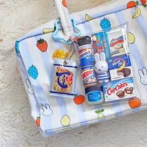 Pu&ograve; includere: Una borsa tote bianca con strisce azzurre e illustrazioni di frutta, dotata di una pochette trasparente fissata da una clip blu a forma di stella. La pochette contiene repliche in miniatura di alimenti, una statuetta Miffy e un vasetto di Vaselina.