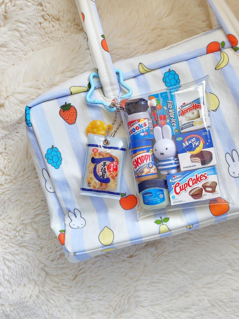Pu&ograve; includere: Una borsa tote bianca con strisce azzurre e illustrazioni di frutta, dotata di una pochette trasparente fissata da una clip blu a forma di stella. La pochette contiene repliche in miniatura di alimenti, una statuetta Miffy e un vasetto di Vaselina.