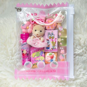 May include: A clear plastic bag filled with pink and white candies, a small bear figurine, and various miniature food items. The bag has a pink zipper and the words "WHITE CHOCOLATE" printed on it.
