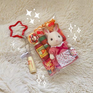 Puede incluir: Una bolsa de pl&aacute;stico transparente contiene una mu&ntilde;eca de conejo blanco con un vestido rosa, alimentos en miniatura y una peque&ntilde;a lata con texto japon&eacute;s. Un llavero rojo en forma de estrella y una botella en miniatura est&aacute;n adjuntos.