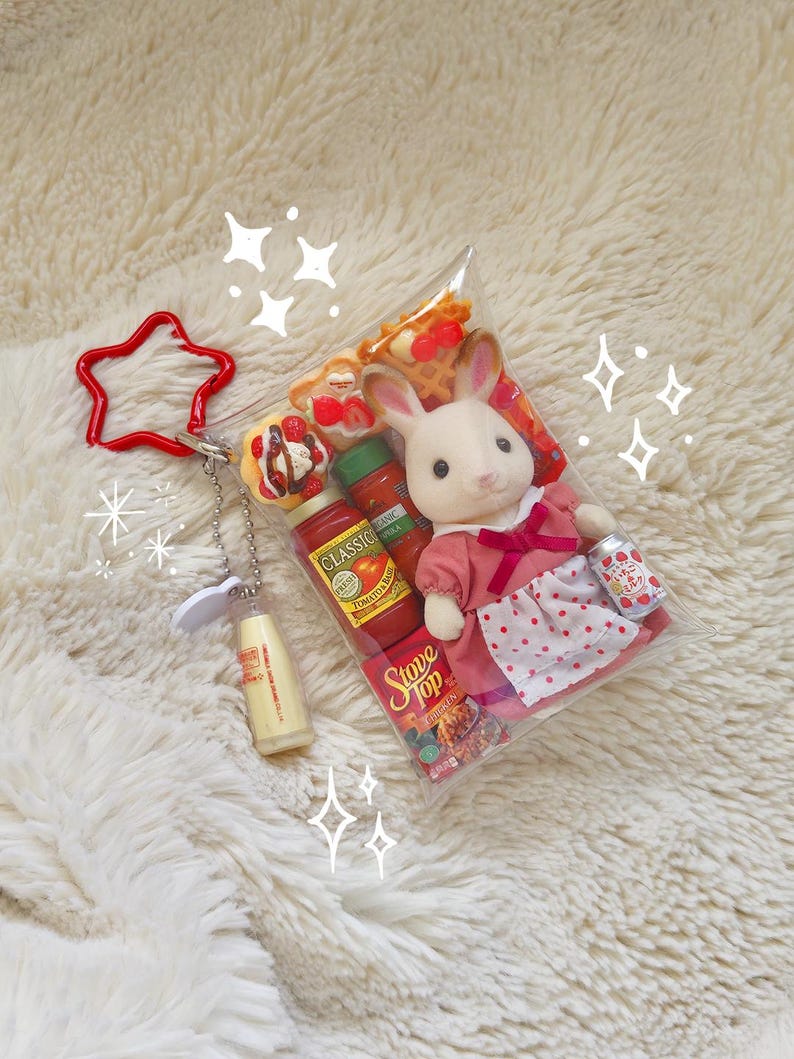 Puede incluir: Una bolsa de pl&aacute;stico transparente contiene una mu&ntilde;eca de conejo blanco con un vestido rosa, alimentos en miniatura y una peque&ntilde;a lata con texto japon&eacute;s. Un llavero rojo en forma de estrella y una botella en miniatura est&aacute;n adjuntos.