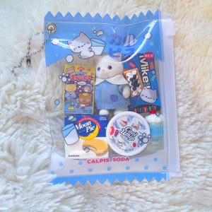 May include: A clear plastic bag with a blue zig-zag top, filled with miniature Japanese snacks and a small white cat doll wearing a blue dress. The bag has a silver chain and the words "CALPIS SODA" printed on it.