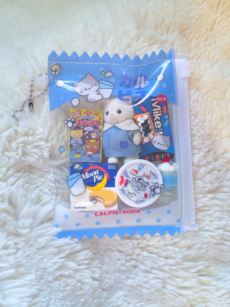 May include: A clear plastic bag with a blue zig-zag top, filled with miniature Japanese snacks and a small white cat doll wearing a blue dress. The bag has a silver chain and the words "CALPIS SODA" printed on it.