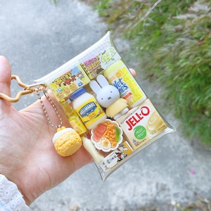 Peut inclure: Une pochette rectangulaire transparente en porte-cl&eacute;s remplie de miniatures alimentaires et de personnages, incluant un pot de mayonnaise Hellmann's Real, une bo&icirc;te de Jell-O vanille, une figurine Miffy, un bol de nouilles et une breloque p&acirc;tisserie jaune.