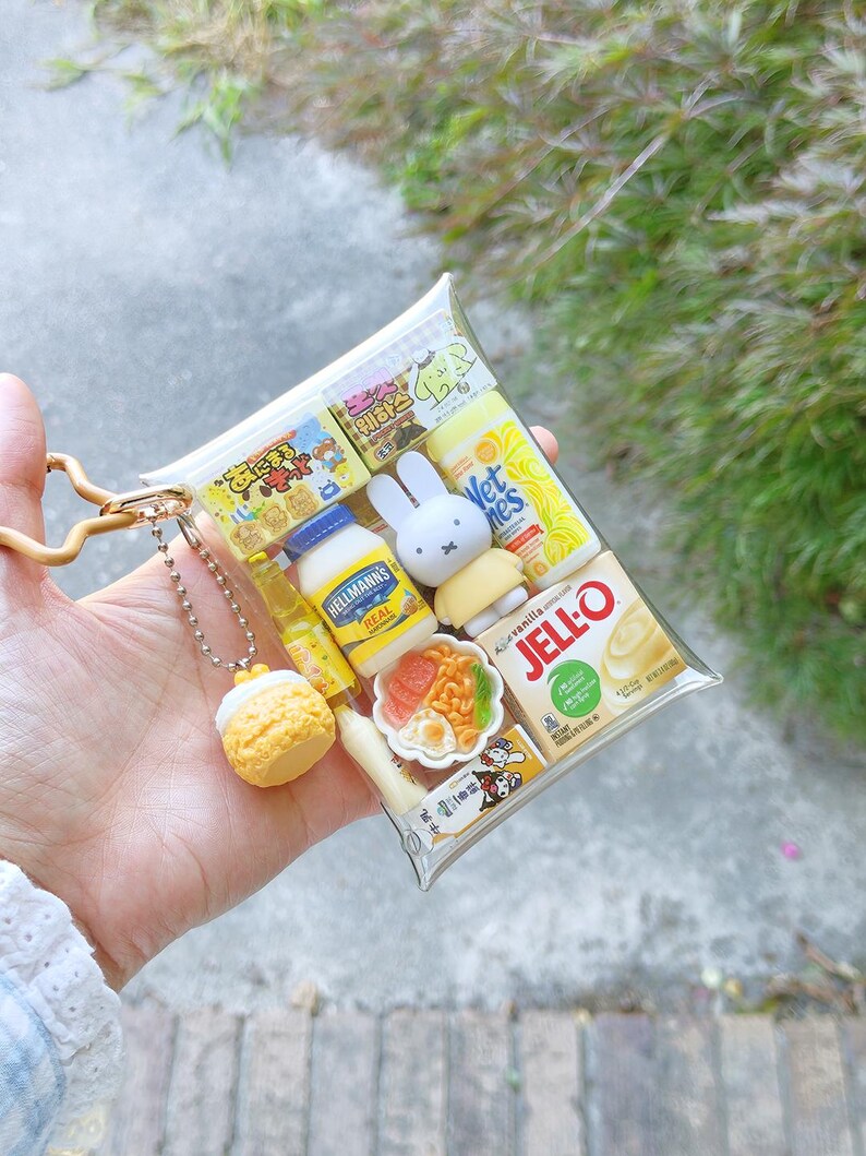 Peut inclure: Une pochette rectangulaire transparente en porte-cl&eacute;s remplie de miniatures alimentaires et de personnages, incluant un pot de mayonnaise Hellmann's Real, une bo&icirc;te de Jell-O vanille, une figurine Miffy, un bol de nouilles et une breloque p&acirc;tisserie jaune.