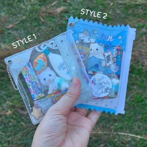 May include: Two clear, zippered pouches filled with miniature items. One pouch, labeled "STYLE 1", contains a white kitten figurine, candy, and a small bottle of Dove. The other, labeled "STYLE 2", has a kitten figurine and various small items. The background is green grass.