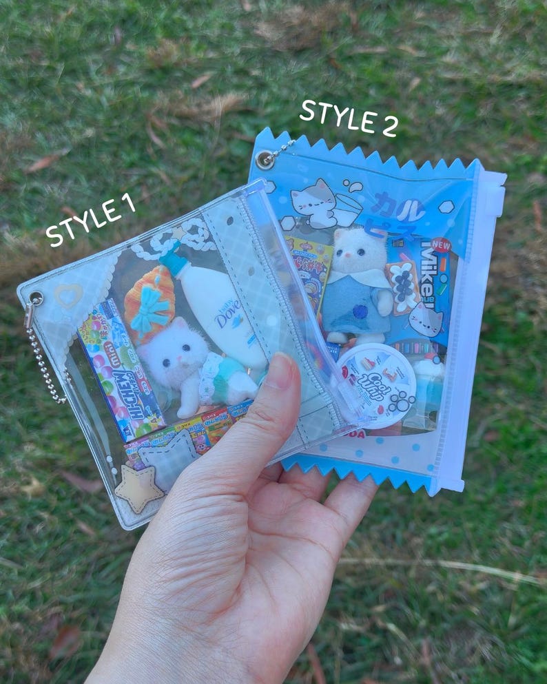 May include: Two clear, zippered pouches filled with miniature items. One pouch, labeled "STYLE 1", contains a white kitten figurine, candy, and a small bottle of Dove. The other, labeled "STYLE 2", has a kitten figurine and various small items. The background is green grass.