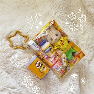 May include: A clear plastic bag filled with miniature food items and a small doll. The bag contains a doll wearing a yellow shirt and green pants, along with small food products like mayonnaise and chips. A gold star-shaped keychain is attached.