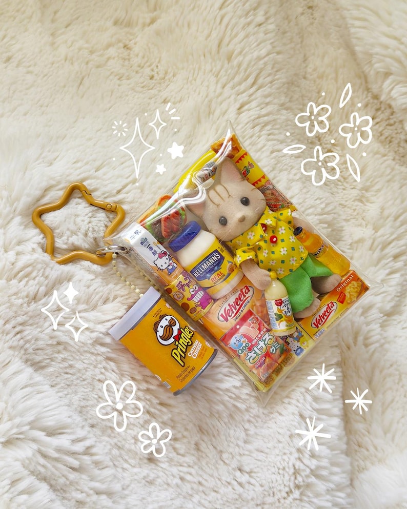 May include: A clear plastic bag filled with miniature food items and a small doll. The bag contains a doll wearing a yellow shirt and green pants, along with small food products like mayonnaise and chips. A gold star-shaped keychain is attached.