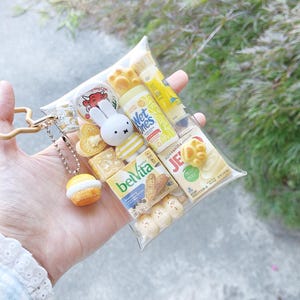 Peut inclure: Une pochette en plastique transparent remplie de r&eacute;pliques miniatures de produits alimentaires et d'hygi&egrave;ne, incluant une figurine Miffy, une bo&icirc;te de biscuits BelVita, une bo&icirc;te Jell-O, un tube Wet Ones et un fromage Laughing Cow, avec un charm cupcake.