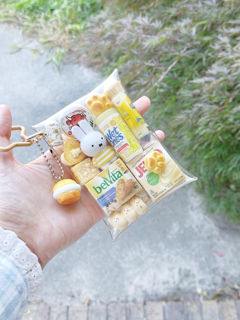 Peut inclure: Une pochette en plastique transparent remplie de r&eacute;pliques miniatures de produits alimentaires et d'hygi&egrave;ne, incluant une figurine Miffy, une bo&icirc;te de biscuits BelVita, une bo&icirc;te Jell-O, un tube Wet Ones et un fromage Laughing Cow, avec un charm cupcake.