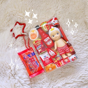 May include: A clear plastic bag filled with miniature food items, a doll, and a red star-shaped keychain. The bag contains small bottles of hot sauce, canned goods, and a doll wearing a white hat and red plaid shorts. The keychain is red.