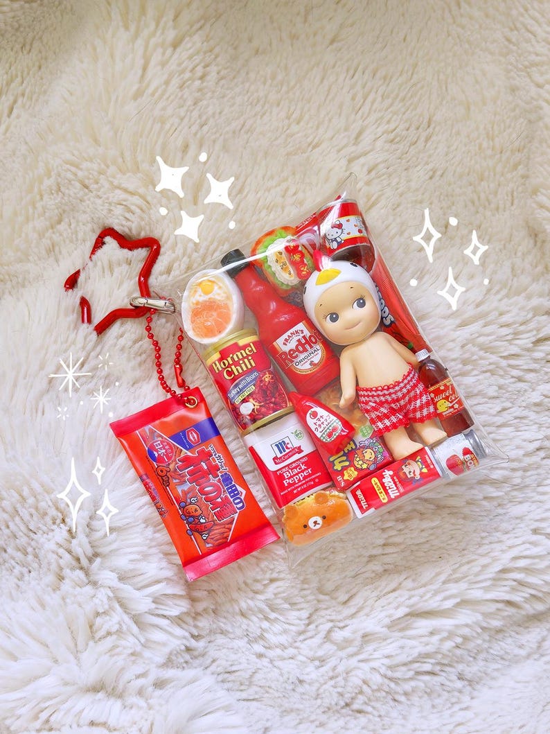 May include: A clear plastic bag filled with miniature food items, a doll, and a red star-shaped keychain. The bag contains small bottles of hot sauce, canned goods, and a doll wearing a white hat and red plaid shorts. The keychain is red.