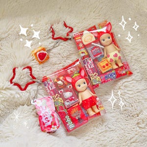 May include: Two small dolls in clear plastic packaging, each wearing strawberry-themed outfits. One doll has a strawberry hat and shorts, the other has a strawberry hat and shorts. The packaging includes miniature snacks and candies. A red star-shaped keychain and a strawberry-shaped keychain are also visible.