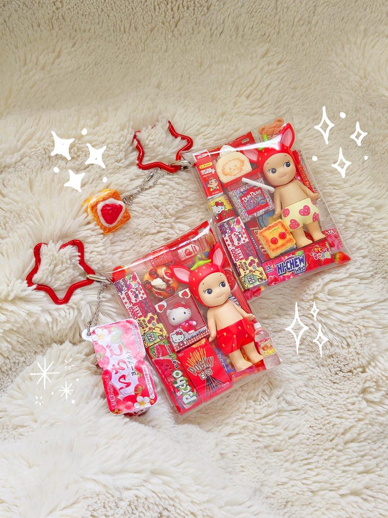 May include: Two small dolls in clear plastic packaging, each wearing strawberry-themed outfits. One doll has a strawberry hat and shorts, the other has a strawberry hat and shorts. The packaging includes miniature snacks and candies. A red star-shaped keychain and a strawberry-shaped keychain are also visible.