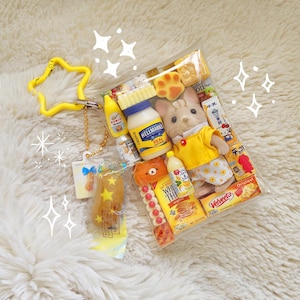 May include: A clear plastic bag filled with miniature food items, a doll, and a yellow star-shaped keychain. The food items include a bottle of Hellmann's Real mayonnaise, a bottle of yellow mustard, and a box of Velveeta cheese.