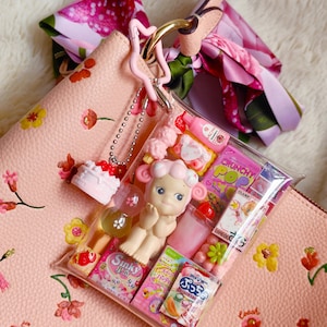 Pu&ograve; includere: Una borsa rosa con un motivo floreale, una sciarpa rosa e un sacchetto di plastica trasparente pieno di caramelle rosa e una bambola. Un piccolo ciondolo a forma di torta &egrave; appeso alla borsa.