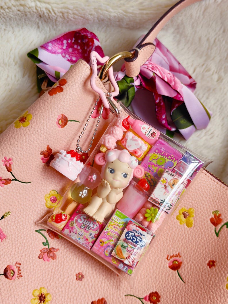 Pu&ograve; includere: Una borsa rosa con un motivo floreale, una sciarpa rosa e un sacchetto di plastica trasparente pieno di caramelle rosa e una bambola. Un piccolo ciondolo a forma di torta &egrave; appeso alla borsa.