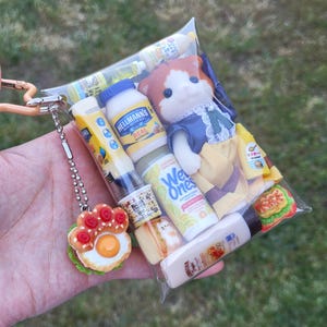 Op de afbeelding: Een doorzichtige plastic zak gevuld met miniatuurvoedsel, een pop en een sleutelhanger in de vorm van een poot. De zak bevat een Hellmann's mayonaisefles, Wet Ones-doekjes en een miniatuursandwich. De sleutelhanger heeft een gebakken ei- en tomatenontwerp.