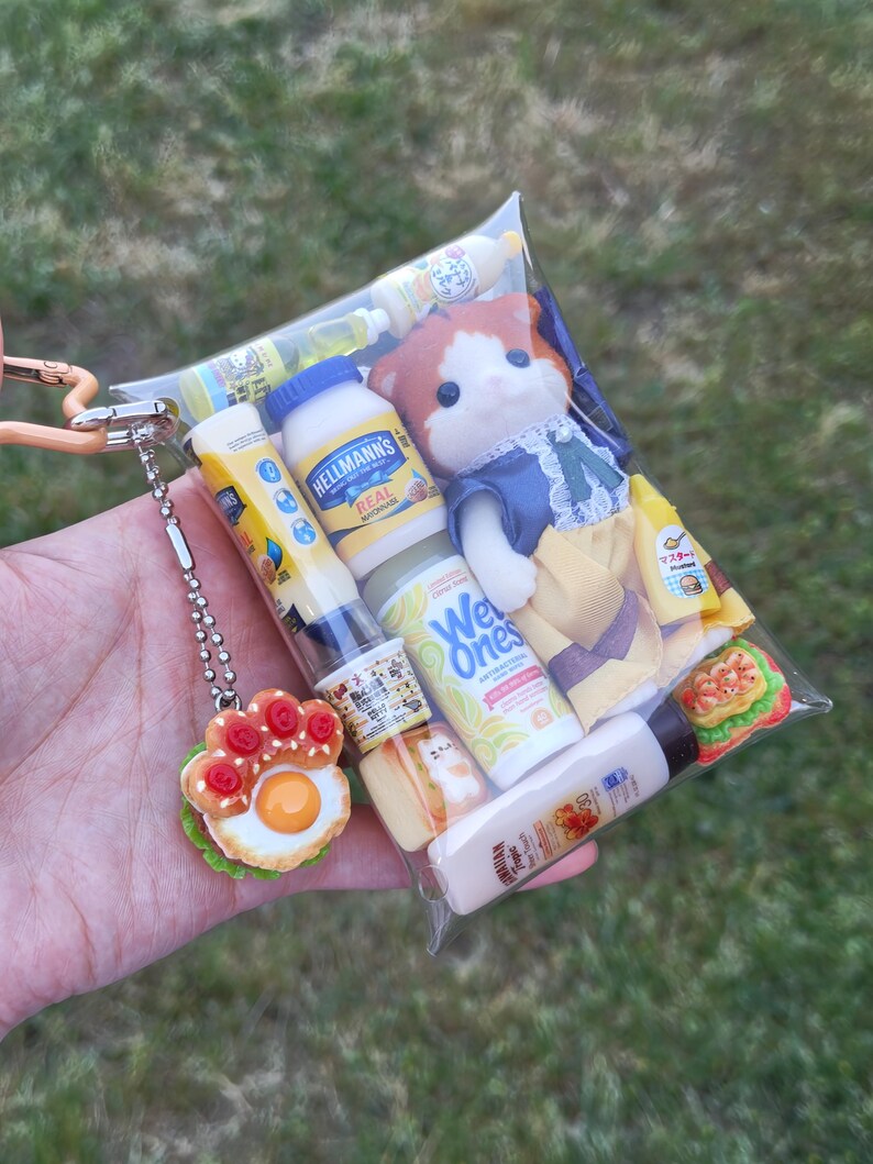 Op de afbeelding: Een doorzichtige plastic zak gevuld met miniatuurvoedsel, een pop en een sleutelhanger in de vorm van een poot. De zak bevat een Hellmann's mayonaisefles, Wet Ones-doekjes en een miniatuursandwich. De sleutelhanger heeft een gebakken ei- en tomatenontwerp.