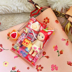 May include: A small doll dressed as a strawberry, packaged with miniature candies and snacks. The doll is in a clear plastic bag, attached to a pink leather bag with a floral pattern. A small, orange, heart-shaped item is also attached.