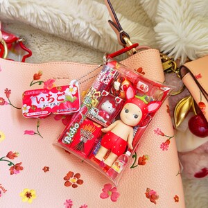 May include: A pink floral handbag with a clear plastic package containing a strawberry-themed doll, Pocky sticks, and other Japanese snacks. A strawberry-shaped keychain and a small yogurt container are also visible.