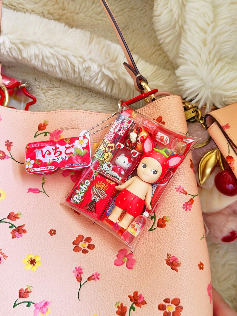 May include: A pink floral handbag with a clear plastic package containing a strawberry-themed doll, Pocky sticks, and other Japanese snacks. A strawberry-shaped keychain and a small yogurt container are also visible.