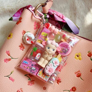 Pu&ograve; includere: Una borsa in pelle rosa con ricami floreali e un cinturino rosa. Un sacchetto di plastica trasparente pieno di giocattoli e caramelle in miniatura &egrave; appeso alla maniglia. Il sacchetto contiene una bambola, un ghiacciolo e altri piccoli oggetti.