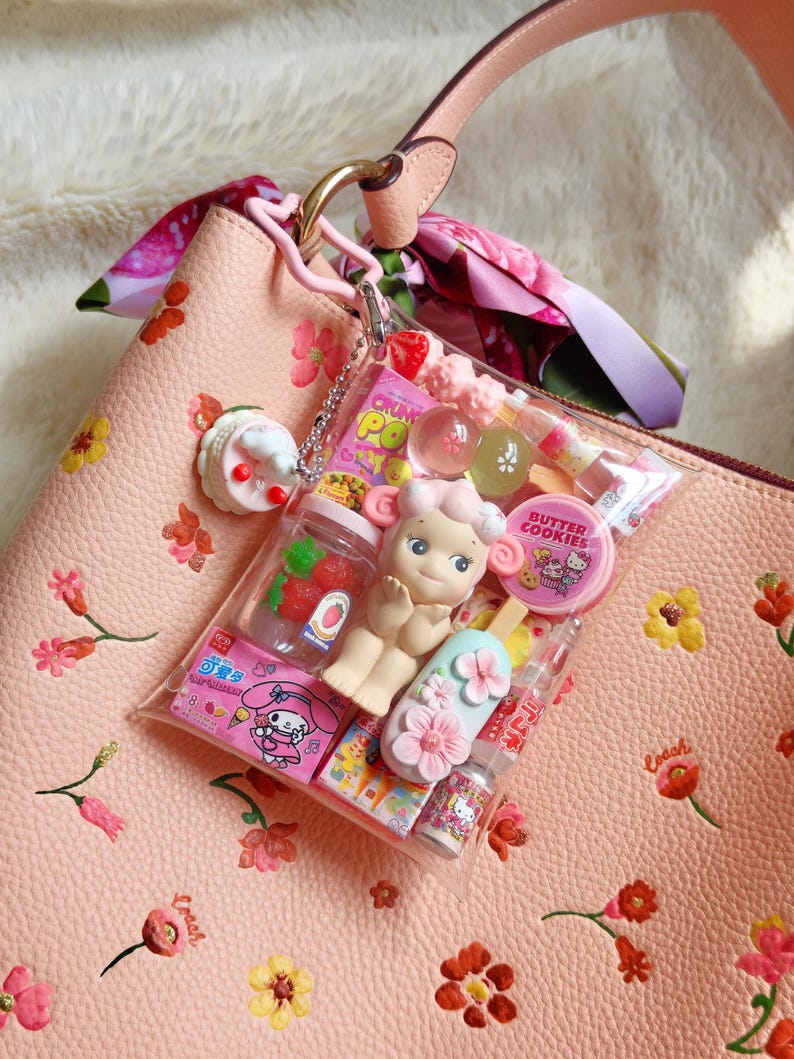 Pu&ograve; includere: Una borsa in pelle rosa con ricami floreali e un cinturino rosa. Un sacchetto di plastica trasparente pieno di giocattoli e caramelle in miniatura &egrave; appeso alla maniglia. Il sacchetto contiene una bambola, un ghiacciolo e altri piccoli oggetti.