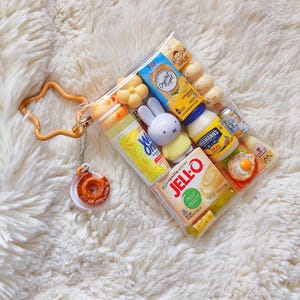 Peut inclure: Une pochette en plastique transparent remplie de miniatures alimentaires et de personnages, dont une bo&icirc;te de Jell-O, de la mayonnaise Hellmann's, du Crystal Light et une figurine Miffy. Un porte-cl&eacute;s orange en forme d'&eacute;toile avec un beignet est attach&eacute;.