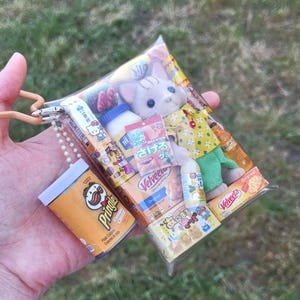 May include: A clear plastic pouch filled with miniature food items and a plush cat toy. The pouch contains small snack boxes, a bottle, and a Pringles can. The cat toy is wearing a yellow shirt and green pants.