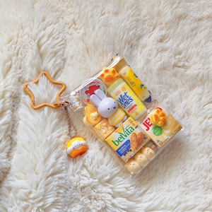 Peut inclure: Une pochette rectangulaire transparente avec un fermoir en forme d'&eacute;toile jaune et une petite breloque suspendue. &Agrave; l'int&eacute;rieur se trouvent des miniatures de produits alimentaires et d'hygi&egrave;ne, dont Belvita, Wet Ones, Jell-O et un lapin.