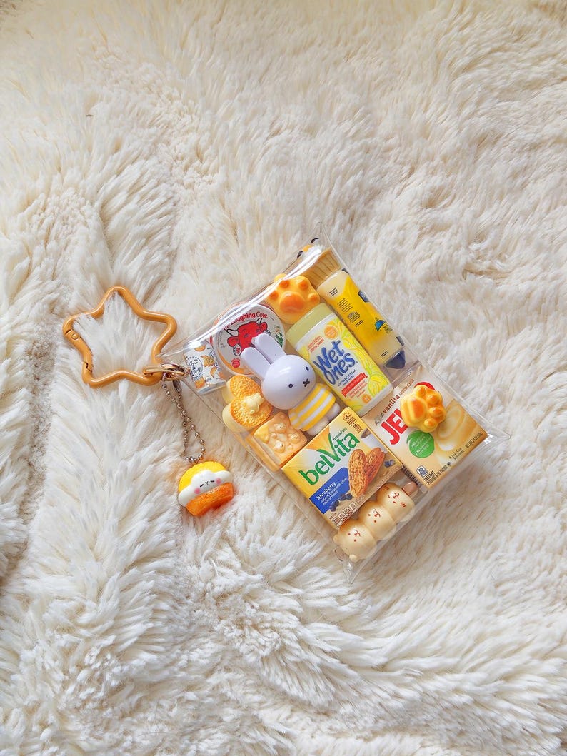 Peut inclure: Une pochette rectangulaire transparente avec un fermoir en forme d'&eacute;toile jaune et une petite breloque suspendue. &Agrave; l'int&eacute;rieur se trouvent des miniatures de produits alimentaires et d'hygi&egrave;ne, dont Belvita, Wet Ones, Jell-O et un lapin.