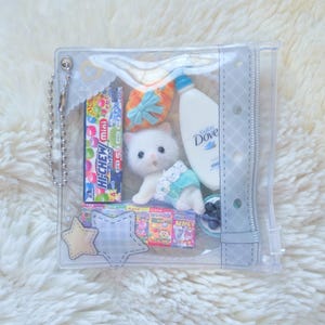 May include: A clear, zippered pouch filled with various items. Includes a plush white kitten, a bottle of Dove baby wash, candy, a small pair of shoes, and star-shaped embellishments. The pouch is on a fluffy white surface.