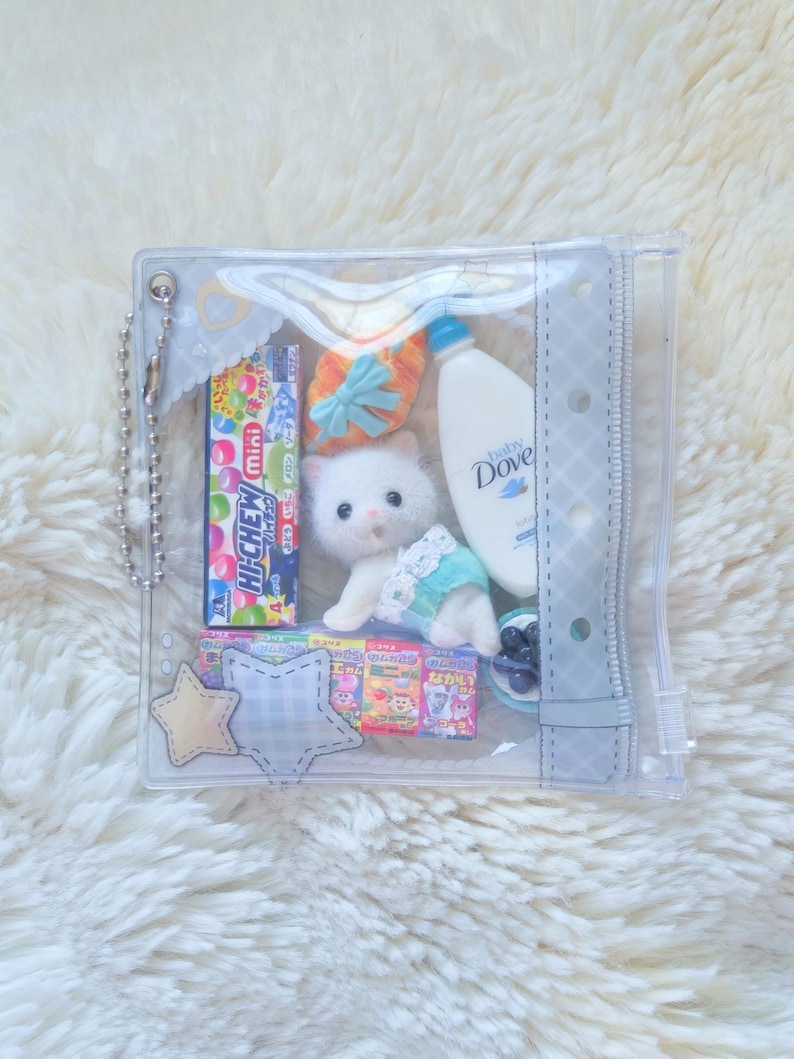 May include: A clear, zippered pouch filled with various items. Includes a plush white kitten, a bottle of Dove baby wash, candy, a small pair of shoes, and star-shaped embellishments. The pouch is on a fluffy white surface.