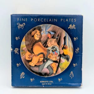 Può includere: Un set di piatti di porcellana pregiata con personaggi de Il Re Leone. Il design del piatto include Simba, Nala, Rafiki e Timon e Pumbaa. La scatola è blu scuro con dettagli dorati e la scritta "FINE PORCELAIN PLATES".