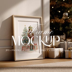 May include: A framed watercolor print featuring a Christmas nutcracker and tree with gifts. The frame is light wood, leaning against a wall. The words "Frame Mockup" are displayed on the frame. Presents and a Christmas tree are in the background.