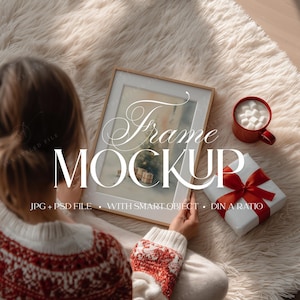 Puede incluir: Una maqueta de arte enmarcada con el texto "Frame Mockup" en un ambiente acogedor. La escena incluye una taza roja con malvaviscos, un regalo envuelto con un lazo rojo y una persona con un suéter rojo y blanco. La imagen está sobre una alfombra esponjosa de color claro.